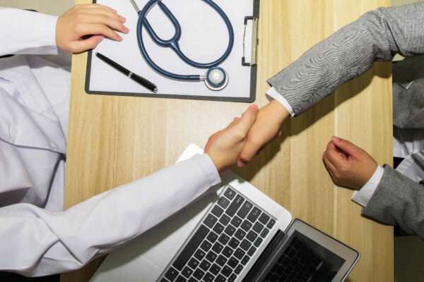 Un médico y un paciente se dan la mano en un escritorio de madera y hablan sobre prevención y tratamiento del Alzheimer. Sobre la mesa hay un portapapeles con un estetoscopio y un bolígrafo, además de una computadora portátil. El médico lleva una bata blanca y el paciente lleva un traje gris.