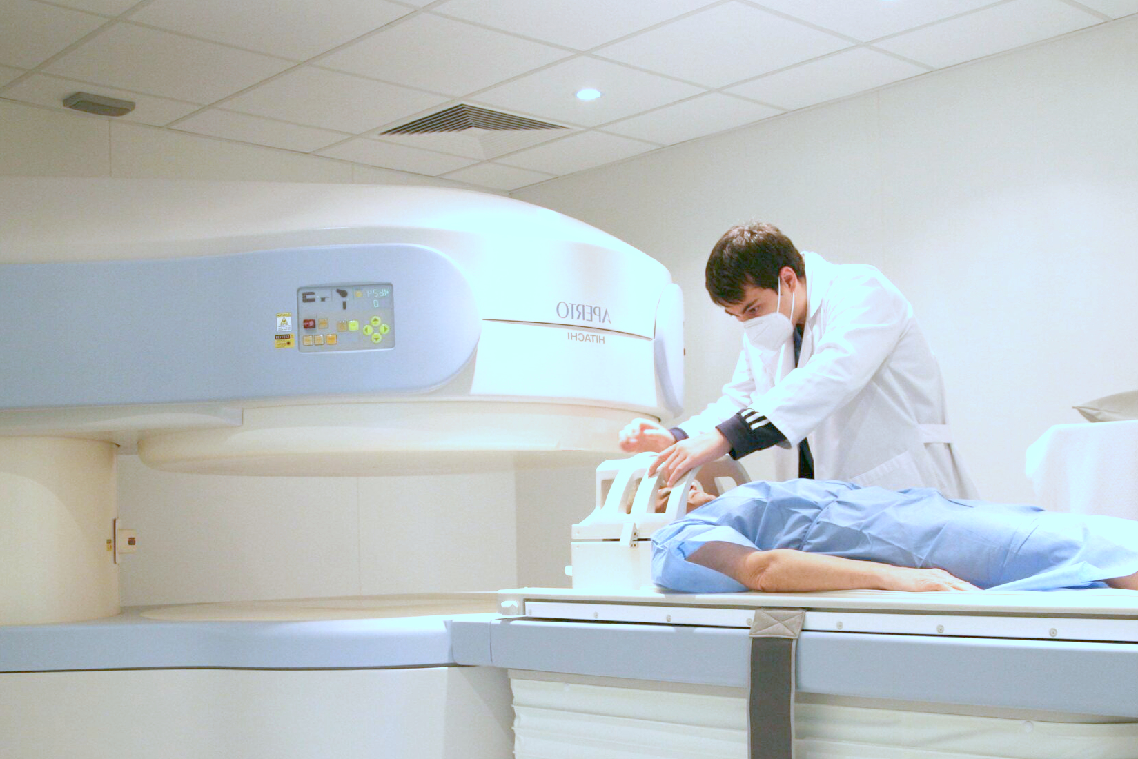 Un técnico con bata blanca ajusta el reposacabezas de un paciente acostado en una máquina de resonancia magnética, lo que subraya el enfoque en la prevención y el tratamiento del Alzheimer. El paciente, con una bata azul, se prepara para la exploración en un entorno clínico con una luz blanca brillante.
