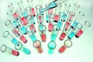 Una vista superior de múltiples tubos de ensayo llenos de líquidos azules y rosados evoca un entorno de laboratorio científico. Una pipeta, suspendida en el aire, sugiere una experimentación precisa en la investigación cardiológica, colocada sobre un fondo claro.