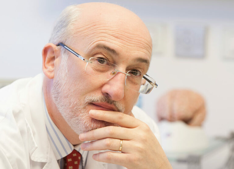 Un hombre calvo, con gafas y barba, vestido con una bata blanca, apoya pensativamente la mano en la barbilla, reflexionando sobre las complejidades de la termodinámica social. En el fondo, un modelo de cerebro humano simboliza la intrincada danza de la entropía sanitaria.