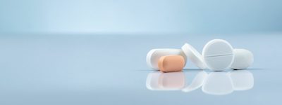 Un primer plano de varias pastillas sobre una superficie lisa y reflectante contra un fondo azul claro resalta los posibles avances del Centro Médico de Neurociencias. Hay cuatro pastillas redondas blancas y una pastilla naranja ovalada, que representan innovaciones en medicina genética.