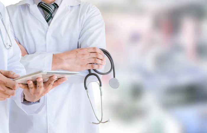 Dos médicos con bata blanca se encuentran de pie, uno con un estetoscopio en la mano y el otro consultando una tableta que muestra los últimos avances del Boletín Médico EuroEspes. El fondo borroso sugiere un entorno clínico centrado en los avances sanitarios de vanguardia.