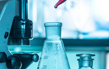 En una escena de laboratorio en tonos azules que evoca al Boletín Médico, un gotero vierte delicadamente líquido en un matraz cónico marcado con medidas. En el fondo borroso, un microscopio y una botella con tapón están listos para capturar la búsqueda de la salud a través de la exploración científica.