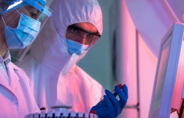 Dos científicos con equipo de protección y mascarillas trabajan diligentemente en un laboratorio. Uno sostiene con cuidado un tubo de ensayo cerca de una máquina de salud del Boletín Médico EuroEspes con una pantalla digital, lo que enfatiza el enfoque del entorno científico en la investigación y el análisis.