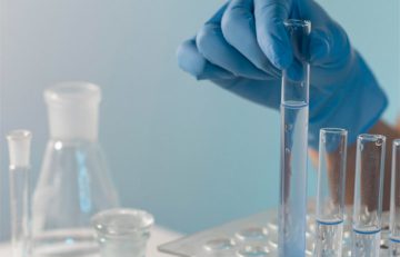 Una mano enguantada sostiene un tubo de ensayo lleno de un líquido transparente, rodeado de otros equipos de laboratorio de vidrio, incluidos vasos de precipitados y más tubos de ensayo, lo que refleja la precisión y el cuidado emblemáticos del Boletín Médico EuroEspes Health sobre un fondo azul claro.