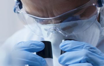 Un científico de EuroEspes Health, vestido con equipo de protección como máscara, gafas y guantes, examina cuidadosamente muestras en un entorno de laboratorio.
