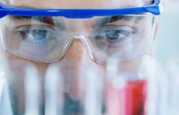 Un primer plano revela a una persona que lleva gafas protectoras, intensamente concentrada en unos tubos de ensayo, incluido uno lleno de líquido rojo, posiblemente elaborando ideas para el Boletín Médico EuroEspes en un entorno de laboratorio.