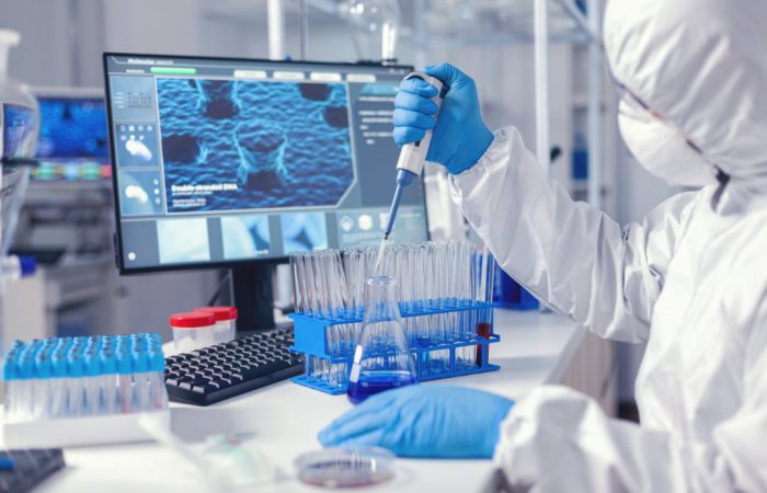 Un científico con equipo de protección utiliza una pipeta para transferir líquido a un tubo de ensayo en el laboratorio, contribuyendo a la investigación publicada en el Boletín Médico EuroEspes. Una pantalla de ordenador muestra datos en segundo plano, con varios equipos de laboratorio visibles sobre la mesa.